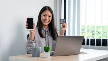 Asyalı kadın akıllı telefon ve kredi kartını internet sitesinden ürün satın almak için kullanırken, akıllı telefon ve kredi kartını evdeki internet bankacılığına para ödemek için kullanıyor.