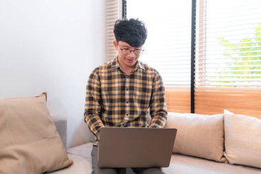 Asyalı adam sosyal medyayı kontrol etmek için dizüstü bilgisayar ekranına bakıyor ve tatilde evdeki oturma odasındaki kanepede oturup sohbet ederken arkadaşlarıyla sohbet ediyor.