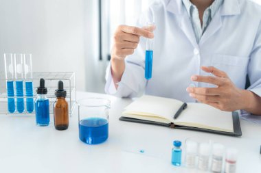 Laboratuvarda kovan 19 aşısı üretmek için mikroskopla çalışırken elinde mavi sıvıyla deney tüpünde test tüpü tutan bilim kadını.