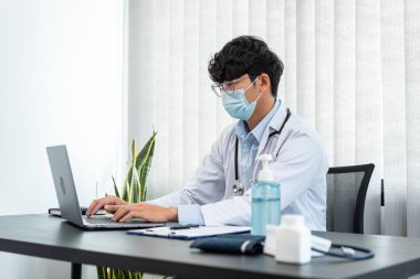 Tıbbi yüz maskesi takan erkek doktor, tedavi semptomları hakkında bilgi aramak ve kliniğinde çalışırken belgeye reçete yazmak için dizüstü bilgisayar kullanıyor.