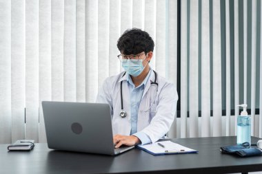 Tıbbi yüz maskesi takan erkek doktor, tedavi semptomları hakkında bilgi aramak ve kliniğinde çalışırken belgeye reçete yazmak için dizüstü bilgisayar kullanıyor.