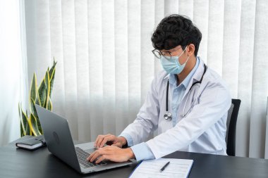 Tıbbi yüz maskesi takan erkek doktor, tedavi semptomları hakkında bilgi aramak ve kliniğinde çalışırken belgeye reçete yazmak için dizüstü bilgisayar kullanıyor.