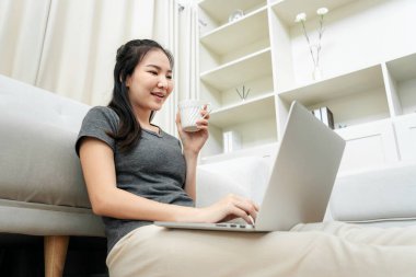 Asyalı serbest çalışan bir kadın kahve fincanı içerken ve dizüstü bilgisayarını kullanarak müşteriden sipariş almak için yerde otururken ürünleri kontrol etmek için merkez ofisteki çevrimiçi pazarlama sırasında...