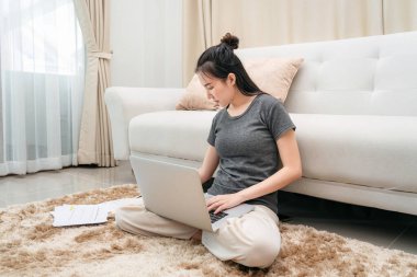 Müşteri siparişi almak için dizüstü bilgisayar kullanan Asyalı serbest kadın ve online pazarlama sırasında ürünleri kontrol ederken yerde oturup müşteri bilgilerini not defterine yazmak