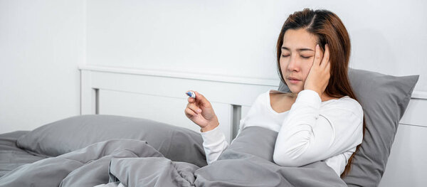 Asian woman feeling sick and headache during using the hand to touching the face and sitting in blanket on the bed while using to checking thermometer to measure body temperature before take medicine 