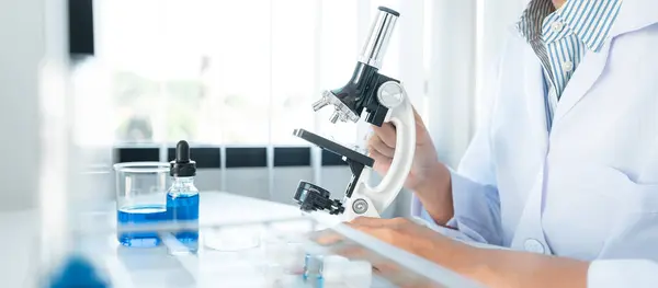 Scientist woman in medical face mask using microscope to research and experiment about coronavirus vaccine while sitting to working in laboratory