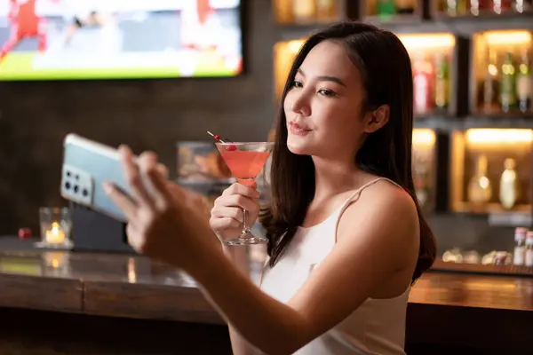 Beautiful woman is holding cocktail and using smartphone to selfie during having enjoy to relaxing while sitting alone to listening music on counter bar in restaurant or hotel bar