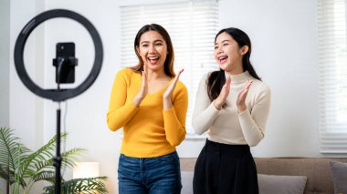 İki kadın kameranın önünde duruyor, gülümsüyor ve alkışlıyorlar. İkisi de sarı gömlek ve siyah pantolon giyiyor. Sahne mutlu ve kutlama dolu.