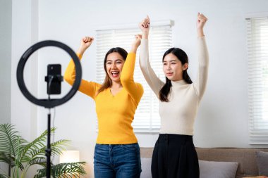 İki kadın kameranın önünde duruyor, biri sarı bir gömlek giyiyor. İkisi de gülümsüyor ve eğleniyor gibi görünüyorlar.