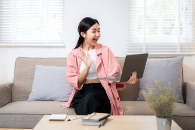 Önünde dizüstü bilgisayarı olan bir kadın kanepede oturuyor. Gülümsüyor ve kameraya el sallıyor. Mutluluk ve bağlantı kavramı