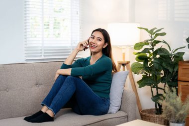Bir kadın kanepede oturuyor ve cep telefonuyla konuşuyor. Gülümsüyor ve sohbetinden zevk alıyor. Oda aydınlık ve rahat bir atmosferi var.