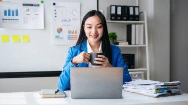 Mavi ceketli bir kadın elinde dizüstü bilgisayar ve bir fincan kahveyle bir masada oturuyor. Gülümsüyor ve işinden zevk alıyor.