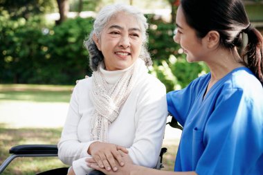Mavi üniformalı bir kadın tekerlekli sandalyedeki yaşlı bir kadına yardım ediyor. Sahne olumlu ve şefkatli.