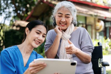 Mavi gömlekli bir kadın tabletli yaşlı bir kadına yardım ediyor.