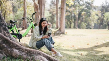Bir kadın yeşil bir bisikletin yanında çimlerin üzerinde oturuyor. Telefonuna bakıyor.