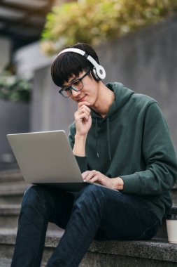 Genç bir adam önünde dizüstü bilgisayarla merdivenlerde oturuyor. Kulaklık ve yeşil bir kapüşonlu giyiyor. Odaklanma ve konsantrasyon kavramı. Adam dizüstü bilgisayarıyla çalışıyor.