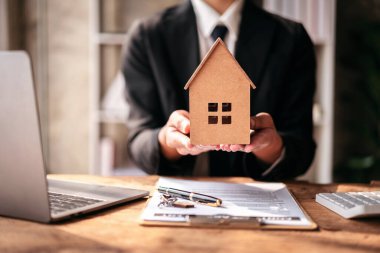 Bir adam model bir evi dizüstü bilgisayarın önünde tutuyor. Takım elbise ve kravat takmış.