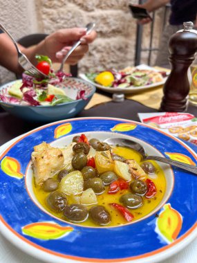 Balık, patates ve zeytinli dekoratif bir tabak. Arka planda taze salata olan eller..