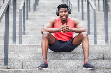 Afro-Amerikan atletik adam kablosuz kulaklıkla müzik dinliyor ve dışarıda gülümsüyor, bir adam merdivenlerde oturuyor ve akıllı bir telefona bakıyor, uzayı kopyalıyor.