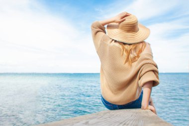 Güneş şapkalı kız denizde iskelede oturur ve ufka bakar, seyahat ve özgürlük konsepti, arka plan