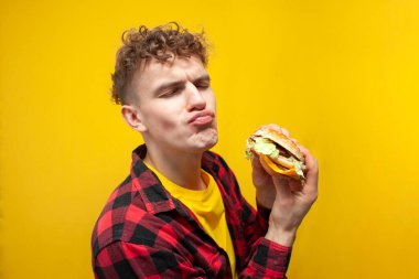 Genç kıvırcık erkek öğrenci sarı arka planda büyük lezzetli hamburgerler yer ve sürpriz, doymuş aç adam fast food yer.