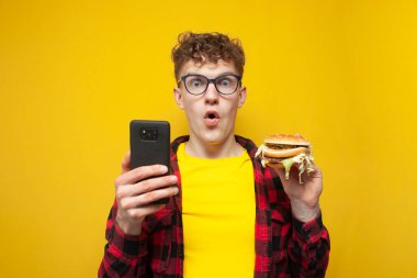 Genç, kıvırcık gözlüklü adam elinde büyük bir hamburger ve akıllı bir telefon tutuyor kameraya şaşkınlıkla bakıyor, telefonu olan bir öğrenci sarı arka planda fast food yiyor.