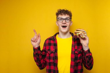 Gözlüklü genç, şaşırmış bir adam hamburger tutuyor ve elini fotokopi makinesinin arka planında gösteriyor. Öğrenci fast food reklamları yapıyor.