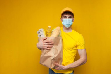 Sarı üniformalı ve tıbbi maskeli genç kurye sarı arka planda bir paket erzak tutuyor. Coronavirus salgınında yemek dağıtan adam.