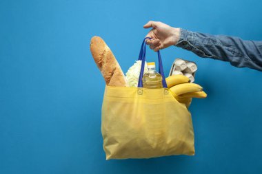 Yiyecek dolu kumaş sarısı eko çanta bir adamın elini mavi arka planda tutar, yemek teslimatı konsepti, plastik reddetme konsepti