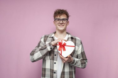 Gözlüklü genç kıvırcık adam pembe arka planda kalp şeklinde bir hediye veriyor. Sevgililer Günü 'nde aşık olan bir adam.
