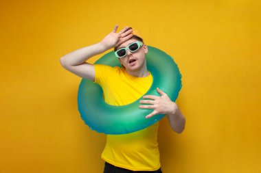 Yaz kıyafetleri ve gözlüklü tatilde olan genç bir adam şişme yüzükle alnını siliyor ve sıcak olduğunu gösteriyor. Yorgun bir adam yaz tatilinde.