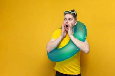 Yazın tatilde olan genç bir adam, şişme yüzme yüzüğü olan bir turist sarı arka planda sürpriz gösteriyor.