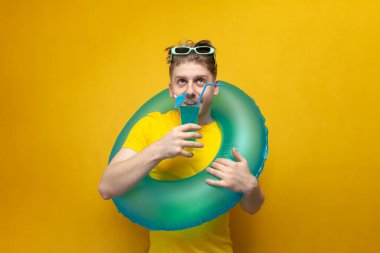 Yaz tatilindeki genç adam sarı arka planda mavi bir kokteyl tutuyor, şişme yüzme çemberi olan bir adam denizde dinleniyor ve ferahlatıcı içecekler içiyor.