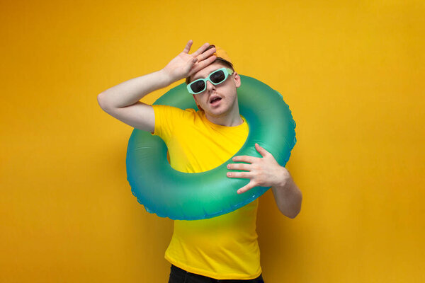 Молодой парень в летней одежде и в очках на отдыхе с надувным кольцом вытирает лоб рукой и показывает, что жарко, уставший мужчина в отпуске летом