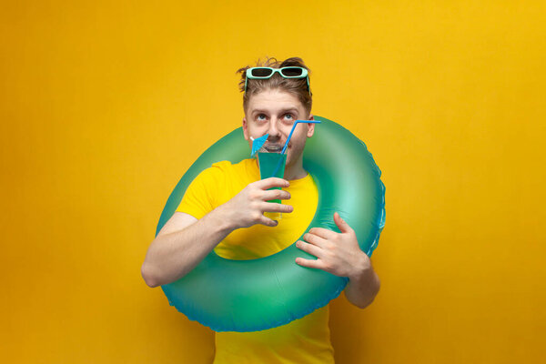 Молодой парень в отпуске летом держит синий коктейль на желтом фоне, мужчина с надувным кругом плавания лежит на море и пьет освежающие напитки