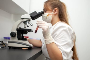 girl doctor in uniform and a medical mask holds test tubes with blood tests and looks through a microscope in the laboratory, a laboratory worker in uniform against the background of the workplace