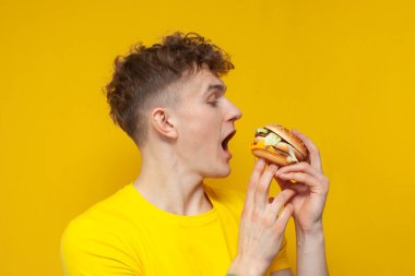 young curly guy holding a burger with his mouth open on a yellow background, a man eats fast food, close-up