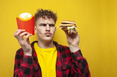 young curly guy with fast food holds french fries and looks suspiciously at a burger, a man chooses between fries and a burger on a yellow background