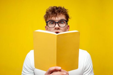 young curly shocked guy in glasses reads a book on a yellow background and shows surprise