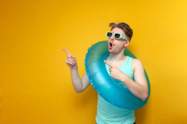 young guy in sunglasses and with inflatable swim ring in the summer on vacation shows surprise and shows with his hands on the copy space space on yellow background