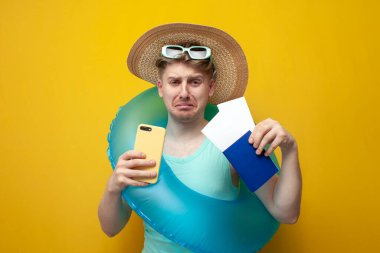 young sad guy in the summer on vacation with a passport and tickets holds a phone and cries on a yellow background, unhappy tourist
