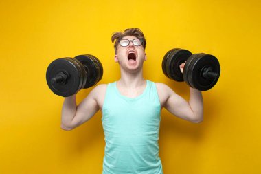 young confident guy student lifts heavy dumbbells on a pink background, motivated nerd in glasses goes in for sports and shouts