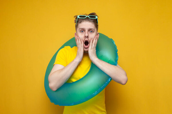 Молодой шокированный парень в отпуске летом с надувным плавательным кольцом показывает сюрприз на желтом фоне