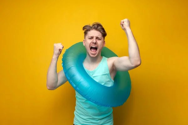 young guy in the summer on vacation with an inflatable swim ring shows a gesture of victory and shouts with raised hands on a yellow background.