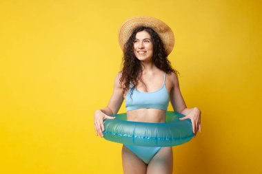 young curly slender brunette girl in a blue swimsuit in summer with an inflatable swimming ring posing on a yellow background and smiling, sexy woman on vacation on the beach looking at the copy space