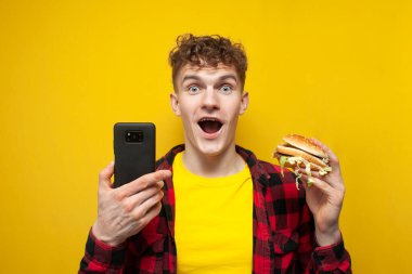 young curly guy holds a big burger and a smartphone and looks at the camera in surprise, a student with a phone eats fast food on a yellow background