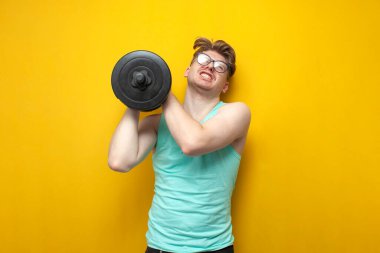 young tired thin guy nerd lifts heavy dumbbells and cries on a yellow background, a confused student in glasses is training