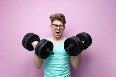 young non-athletic guy in glasses holds heavy dumbbells on a pink background and trains, a serious motivated nerd goes in for sports and screaming