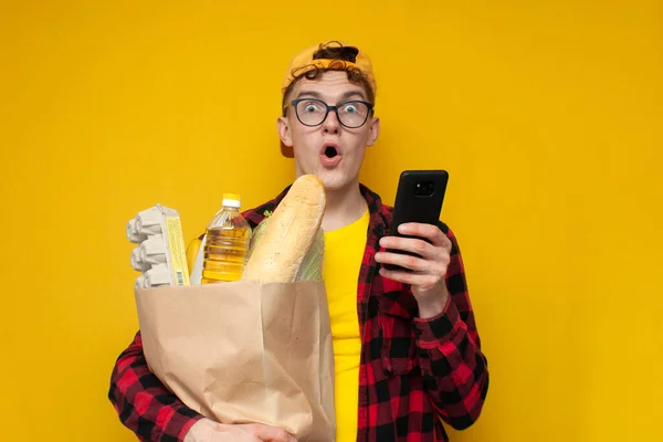 young shocked customer with a package of groceries uses a phone on a yellow background and is surprised, a consumer with food looks into a smartphone and orders food