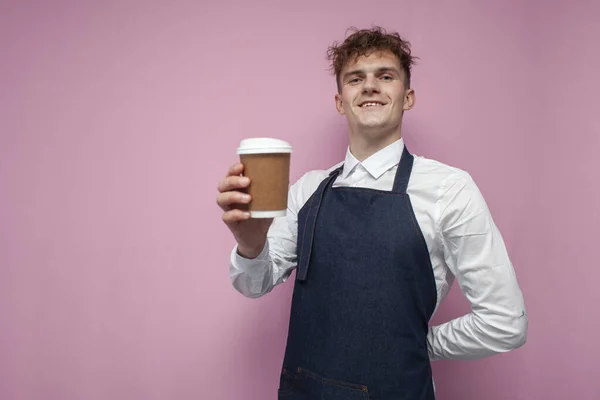 young barista in a white shirt and apron holds a cup of coffee and smiles on a pink background, portrait of a waiter worker guy
